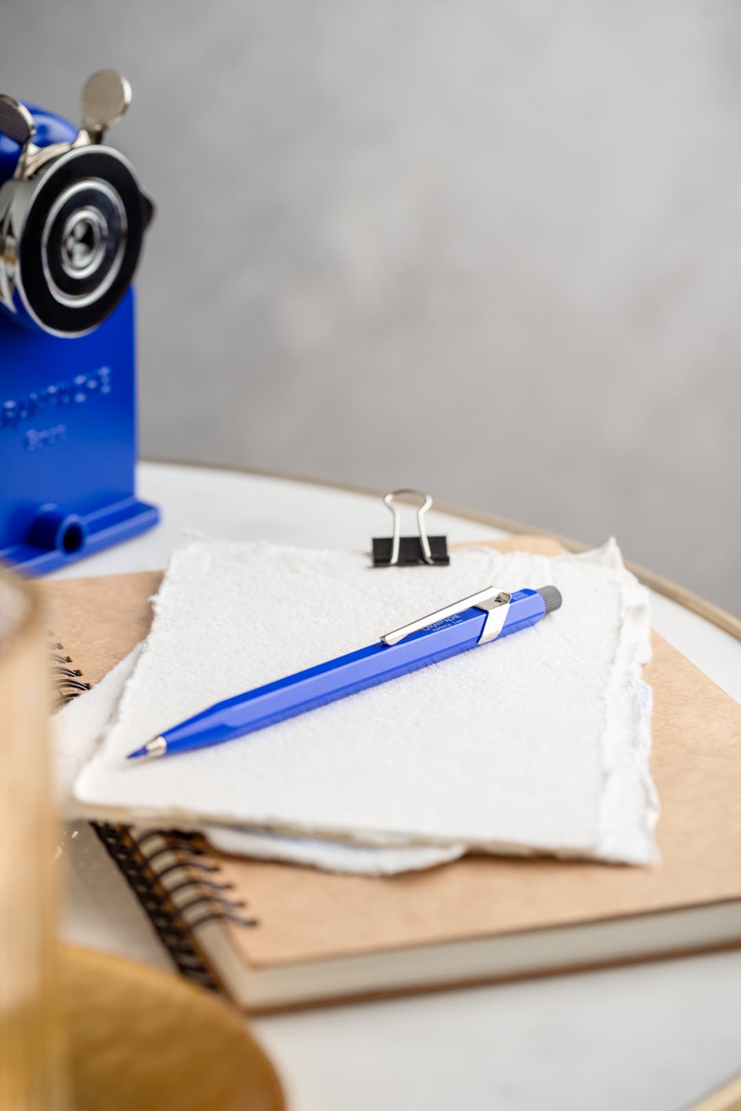 Stylo Caran d'Ache bleu Klein Blue posé sur un carnet blanc avec pince métallique, à côté d'une perforatrice bleue