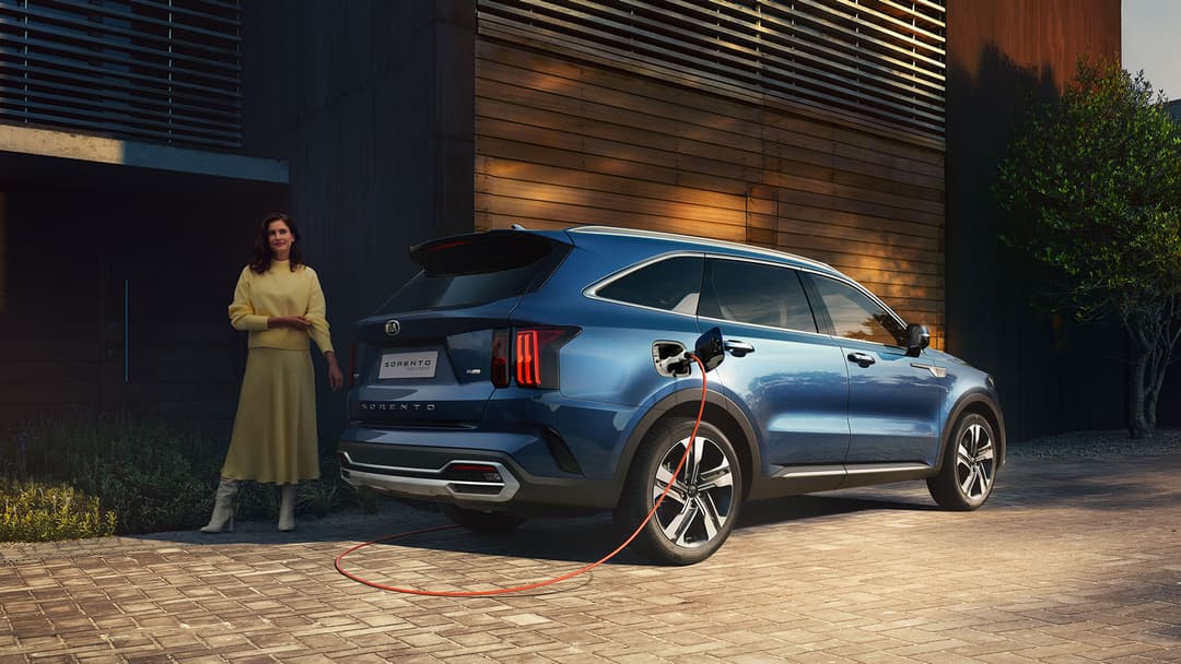 Kia Sorento bleu métallisé en recharge, femme en robe jaune à côté du véhicule hybride devant une façade en bois