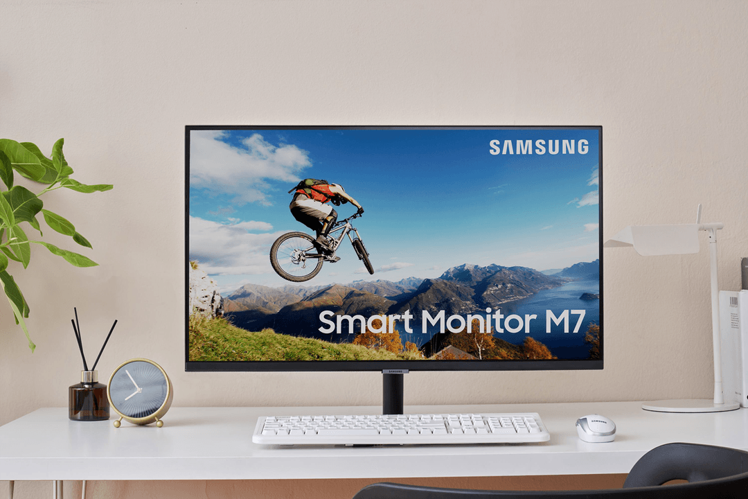 Smart Monitor M7 de Samsung affichant un cycliste en vol sur un paysage montagneux, posé sur un bureau avec clavier et souris