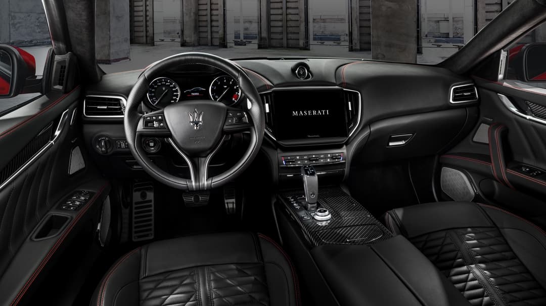Intérieur d'une Maserati 2021 avec tableau de bord noir, volant à trois branches, écran central affichant le logo Maserati et sièges en cuir noir et rouge