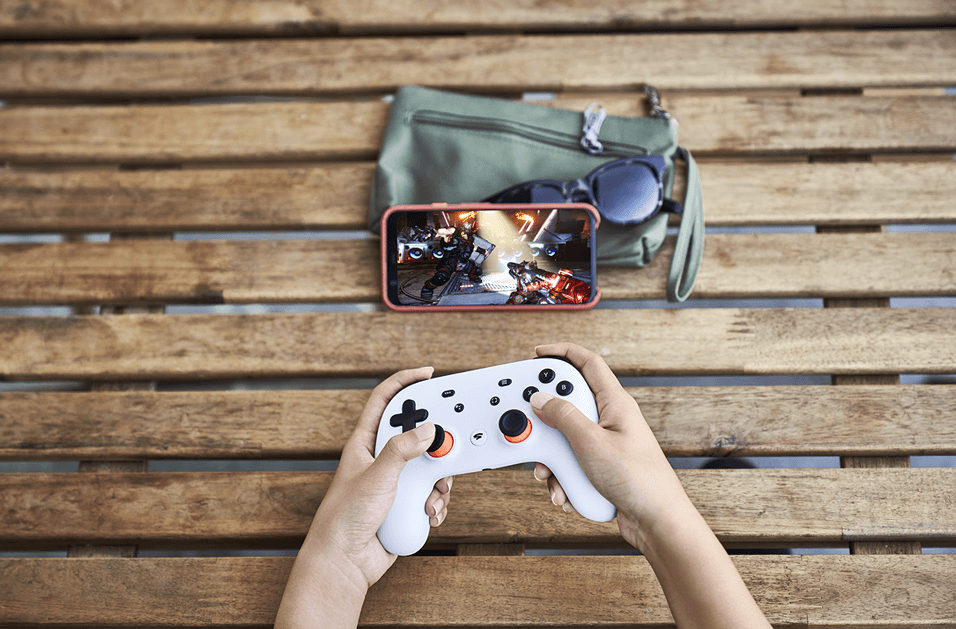 Mains tenant une manette Stadia blanche sur un banc en bois, smartphone rouge affichant un jeu et sac vert à proximité
