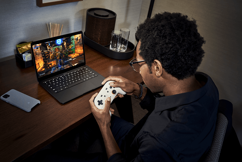 Personne jouant à un jeu vidéo sur un laptop noir avec une manette de contrôle blanche, assise à un bureau en bois