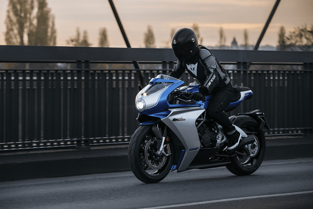 Motocycliste en tenue de course noire sur une MV Agusta blanche et bleue Alpine, circulant sur un pont avec barrière métallique