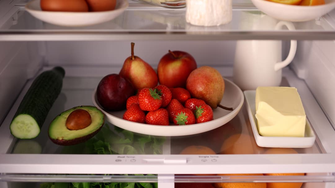 Intérieur de réfrigérateur avec fruits et légumes rangés dans des bols blancs : fraises, pommes, poires, avocat, concombre et beurre