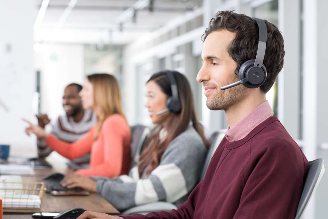 Casque Logitech Zone Wireless gris porté par un homme dans un bureau avec collègues en arrière-plan
