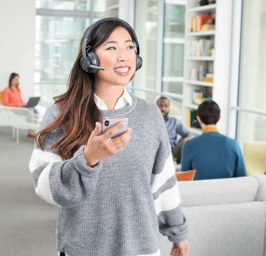Femme portant un casque Logitech Zone Wireless gris avec microphone, tenant un téléphone dans un bureau