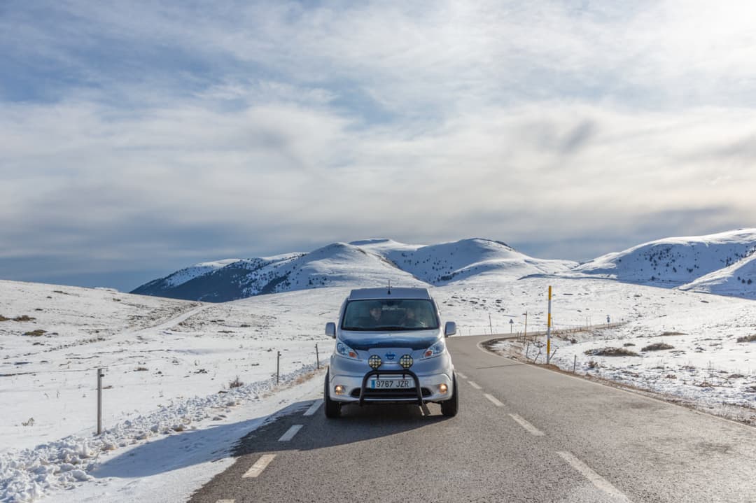 Nissan e-NV200 Winter Camper blanc sur route enneigée en montagne, équipé de barres de toit et accessoires d'hiver