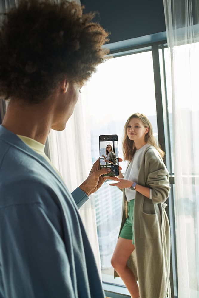 Deux personnes se prenant en photo avec un Samsung Galaxy S21, l'une tenant le téléphone pour photographier l'autre près d'une baie vitrée