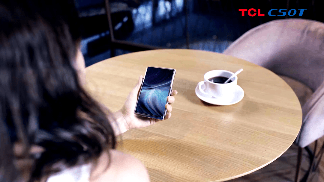 Smartphone TCL CSOT à écran flexible enroulé tenu en main au-dessus d'une table en bois
