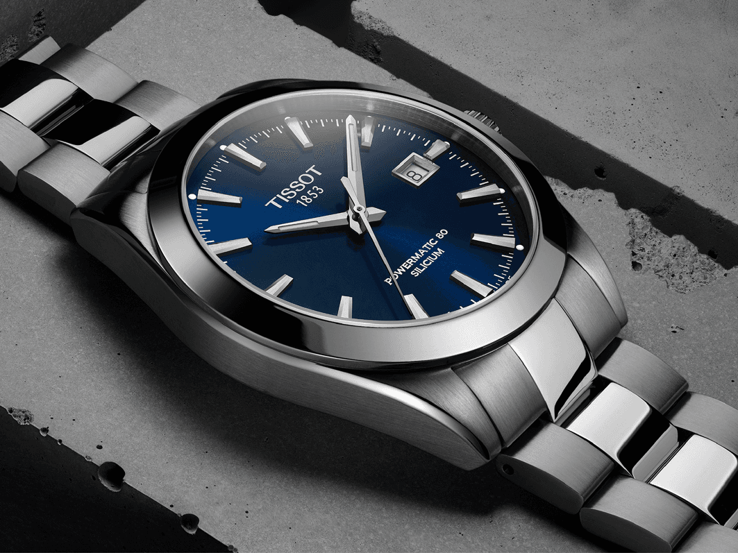 Montre Tissot PRX acier avec cadran bleu marine, bracelet métallique, posée sur surface grise