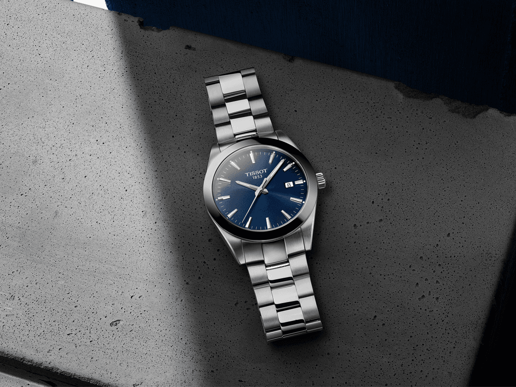 Montre Tissot Gentleman à boîtier et bracelet en acier, cadran bleu foncé et aiguilles dorées, posée sur une surface grise