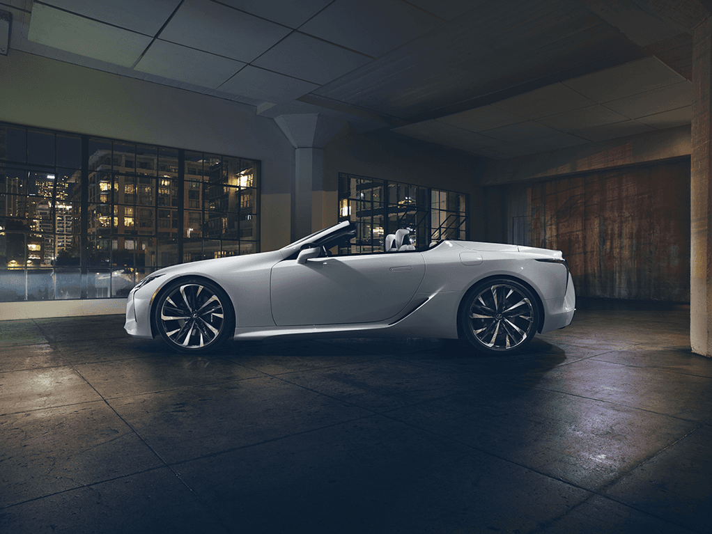 Lexus LC 500 Cabriolet blanc décapotable en profil, stationnée dans un garage moderne avec vue sur une skyline urbaine illuminée