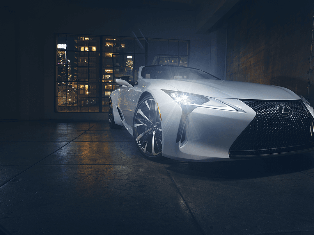 Lexus LC 500 Cabriolet blanc avec toit ouvert, de profil, devant un immeuble illuminé la nuit