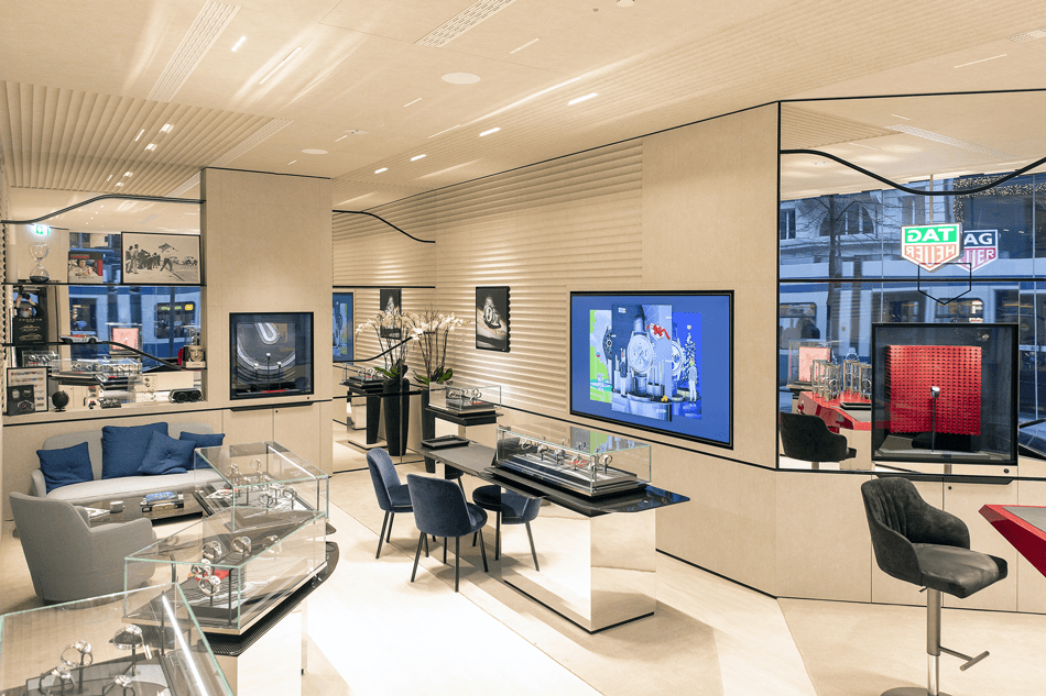 Intérieur épuré de la boutique flagship Tag Heuer à Zurich avec vitrines de montres, écrans vidéo et mobilier design aux teintes beige et bleu