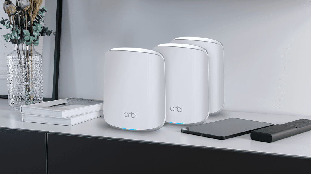Trois routeurs Netgear Orbi blanc posés sur un bureau moderne à côté de livres et d'une tablette