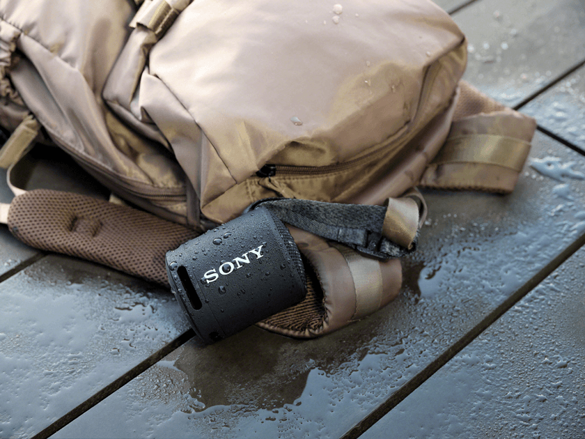 Enceinte Sony SRS-XB13 noire avec dragonne, posée près d'un sac à dos beige sur une surface mouillée