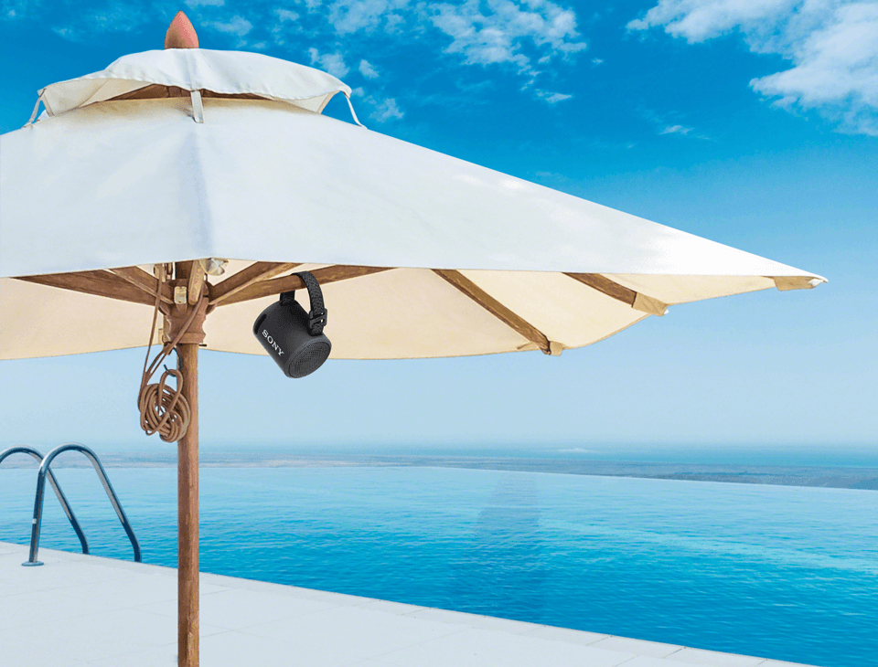 Enceinte Sony SRS-XB13 noire suspendue sous un parasol blanc au bord d'une piscine avec vue sur la mer