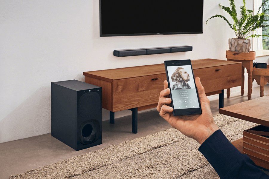 Système home cinéma Sony HT-S40R composé d'une barre soundbar noire, d'un caisson de basses noir et d'un smartphone affichant une application de contrôle, dans un salon