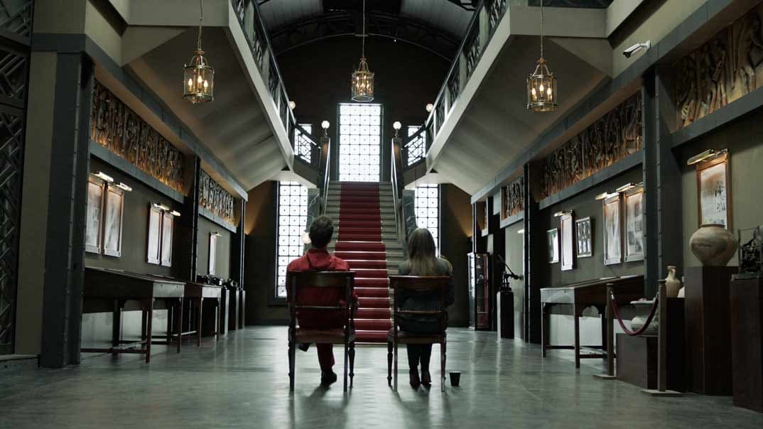 Deux personnages assis face à un escalier rouge dans un musée au décor imposant aux murs noirs et lustres dorés
