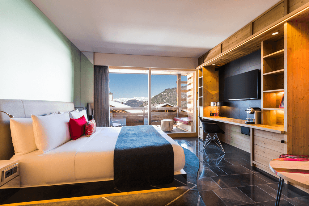 Chambre rénovée du W Verbier avec lit blanc et rouge fluo, mobilier en bois clair, vue panoramique sur les Alpes enneigées