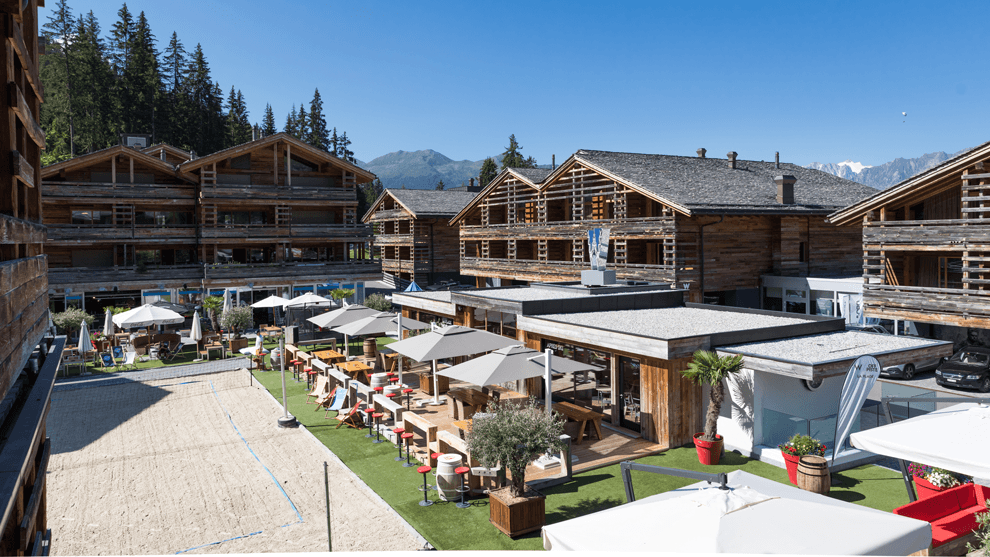 Plage du W Verbier avec terrasse herbeuse, parasols blancs, bâtiments en bois et montagnes enneigées en arrière-plan