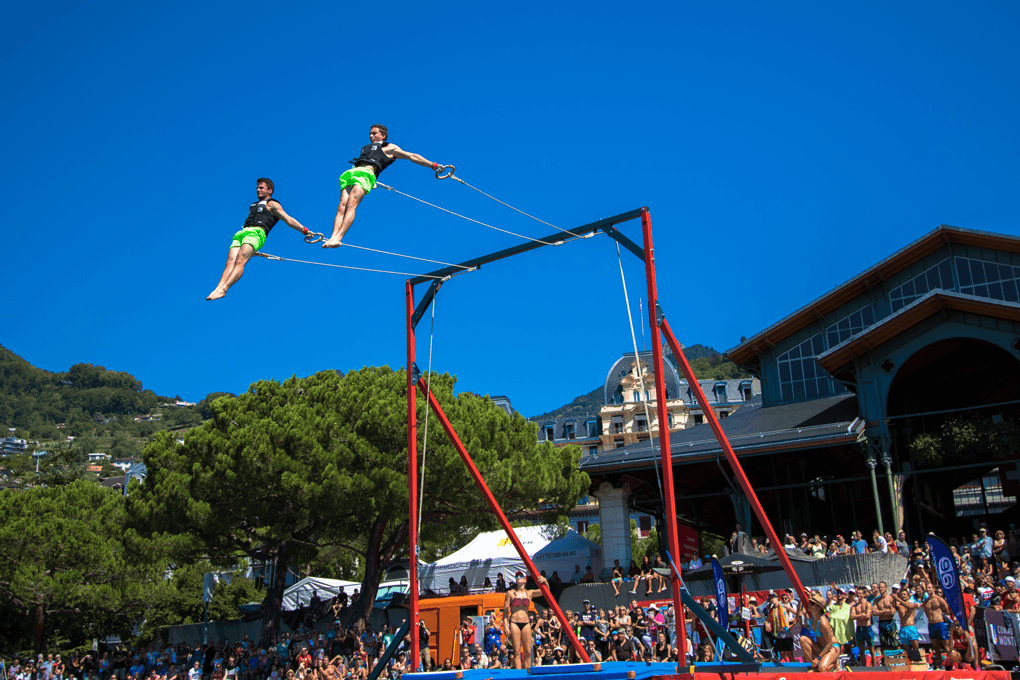 Deux athlètes en short vert fluo effectuent un saut acrobatique suspendu à des cordes au-dessus d'une structure métallique rouge, devant une foule nombreuse sous un ciel bleu