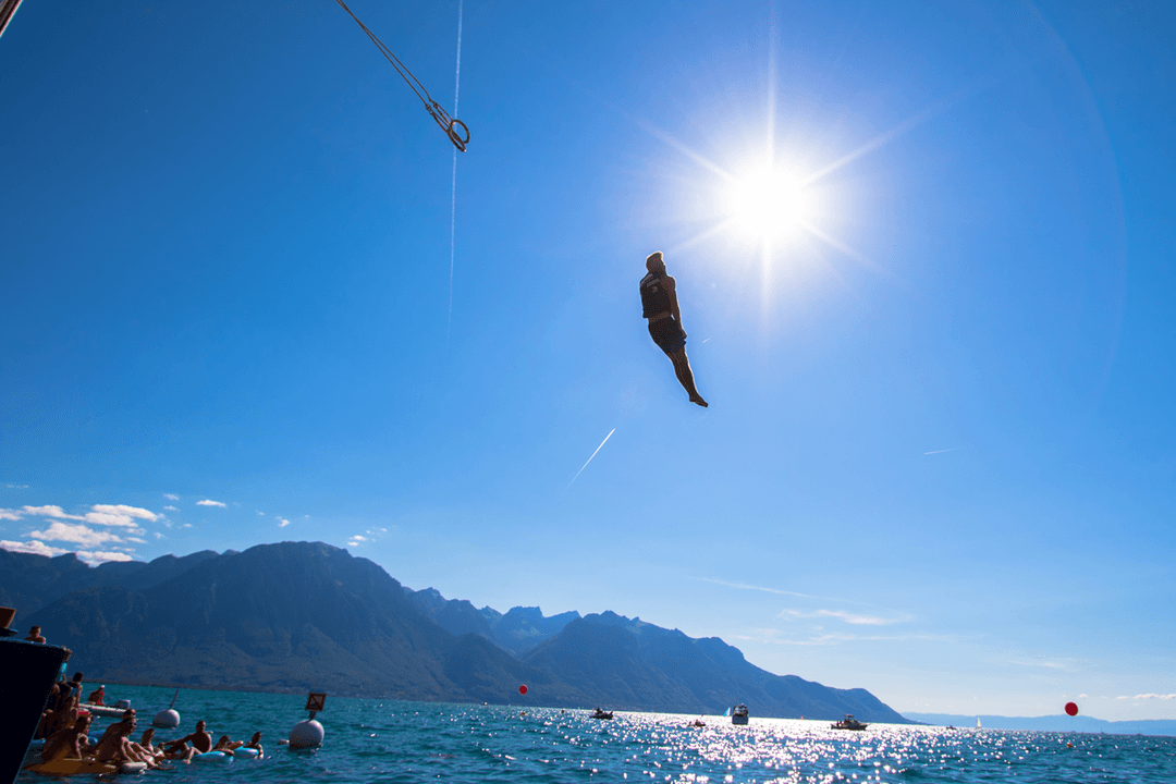 Personne en suspension à la corde au-dessus de l'eau turquoise avec des spectateurs en bord de lac et montagnes en arrière-plan