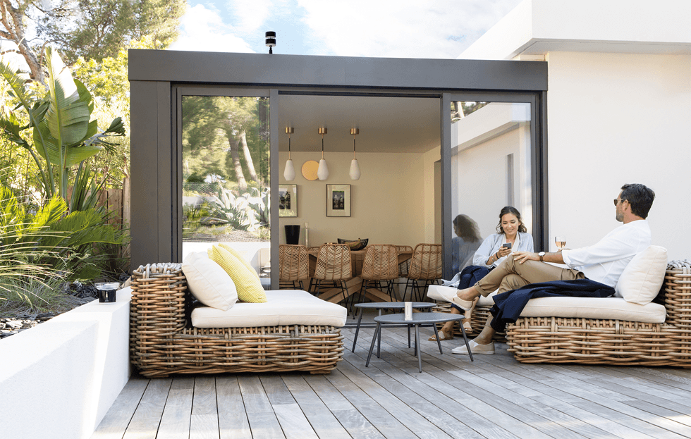 Terrasse avec mobilier en rotin, deux personnes discutent près d'une maison moderne aux grandes baies vitrées, végétation verte en arrière-plan