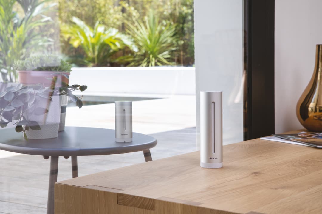 Station météo intelligente Netatmo blanche placée sur un bureau en bois clair, avec un espace de travail lumineux et des plantes en arrière-plan