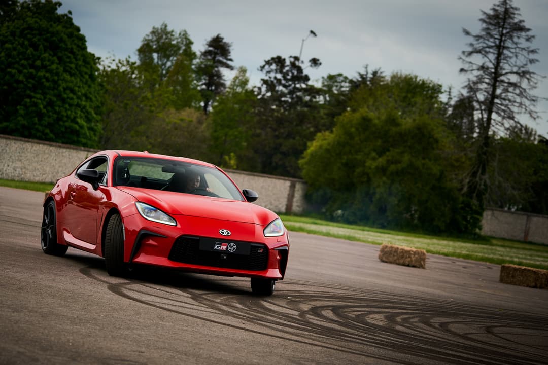 Toyota GR 86 rouge en trois-quarts avant sur un circuit, avec traces de pneus au sol et arbres en arrière-plan