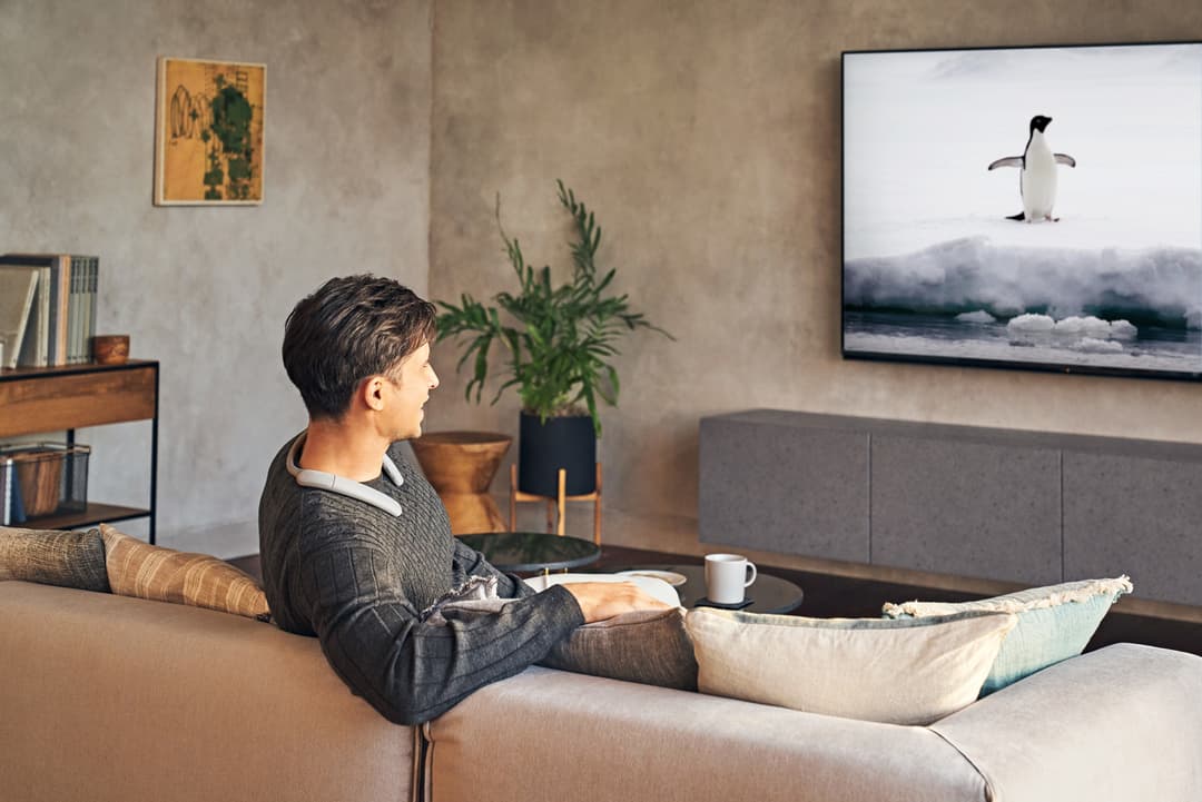 Homme assis sur un canapé regardant la télévision dans un salon moderne, portant une enceinte Bluetooth autour du cou