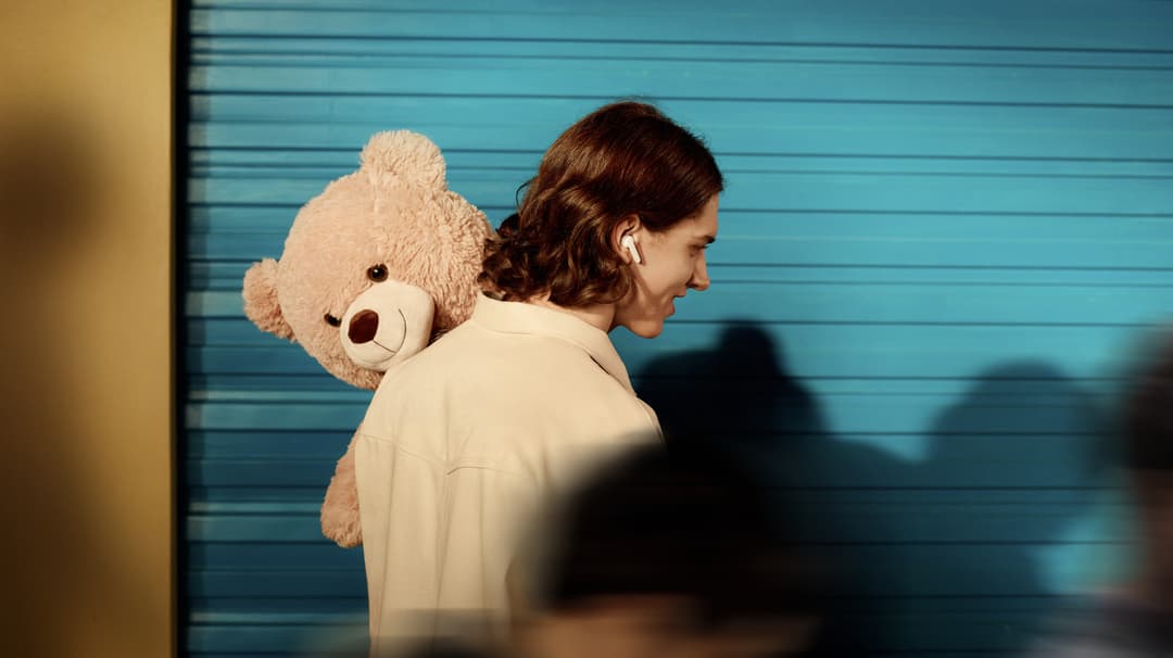 Femme portant des écouteurs blancs, tenant un nounours beige sur son épaule, devant un mur bleu