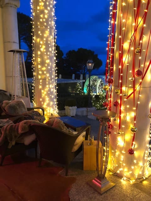 Terrasse illuminée de guirlandes blanches et rouges au crépuscule, avec fauteuils confortables et vue sur le lac