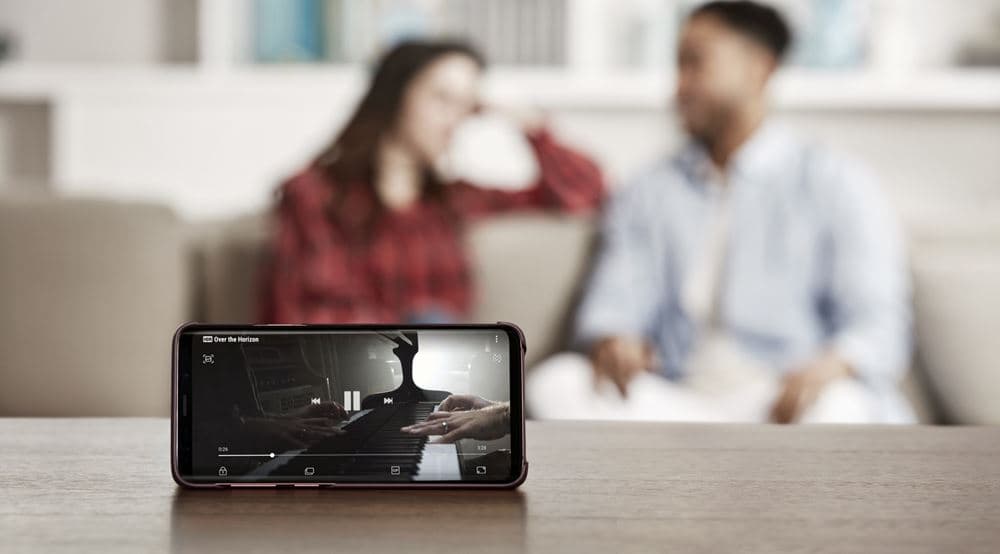Galaxy S9 posé sur une table affichant une vidéo musicale, avec un couple assis sur un canapé en arrière-plan