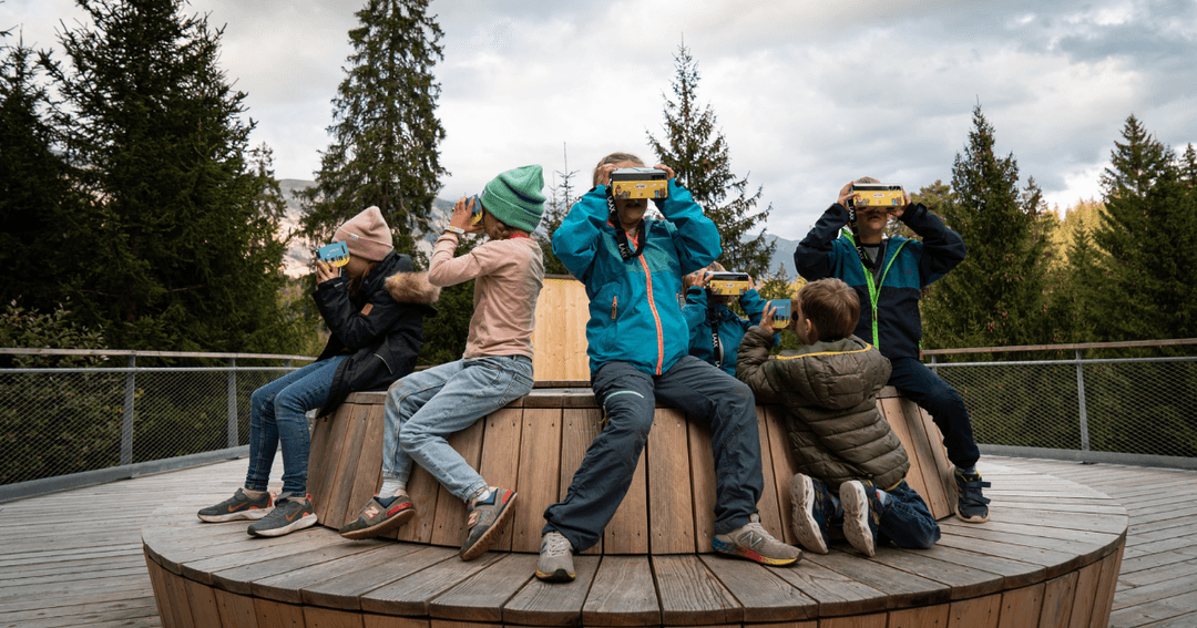 Enfants portant des lunettes de réalité virtuelle Cardboard assis sur une plateforme en bois face aux montagnes des Grisons