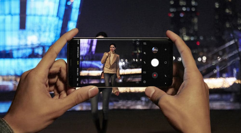 Deux mains tenant un Galaxy S9 qui photographie une femme de nuit en milieu urbain avec éclairages bleus et blancs flous en arrière-plan