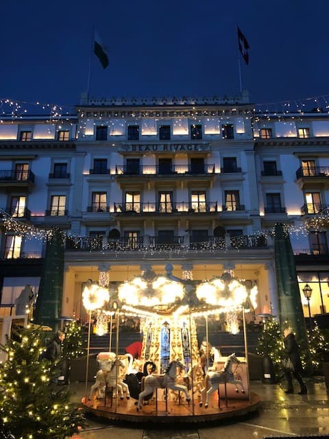 Façade illuminée du Beau-Rivage Palace la nuit, décorée de guirlandes lumineuses, avec un salon de thé en vitrine au rez-de-chaussée