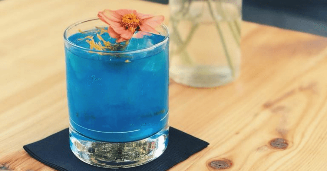 Cocktail bleu vif garni d'une fleur rose, servi sur une table en bois à la Plage à Carouge