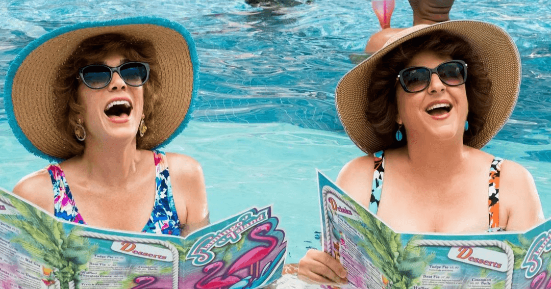 Deux femmes rient en lisant des magazines à la piscine, portant chapeaux de paille et lunettes de soleil