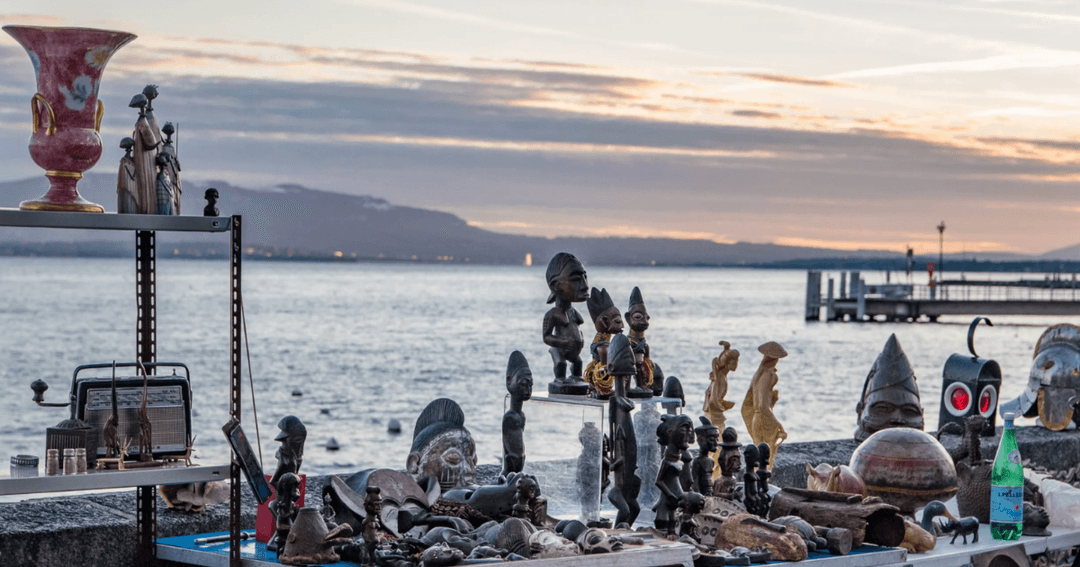 Marché aux puces au bord du Léman à Nyon avec statues, vases et objets de collection disposés sur la rive au coucher du soleil