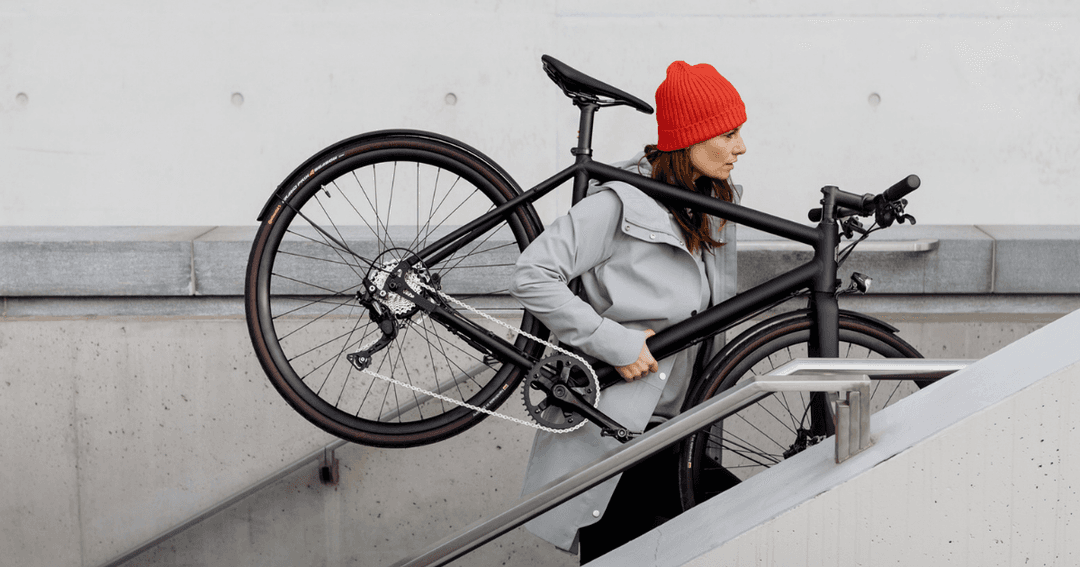 Une femme en bonnet rouge soulève un vélo électrique noir au-dessus de son épaule, devant un mur en béton