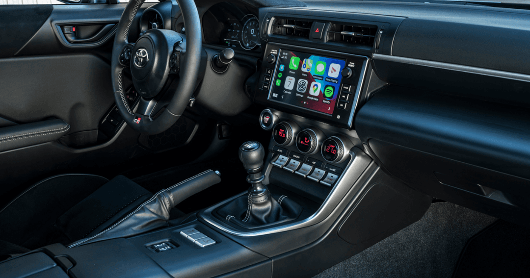 Habitacle de la Toyota GR86 avec volant noir, écran tactile intégrant Apple CarPlay, sièges cuir noir avec surpiqûres argentées et tapis gris foncé