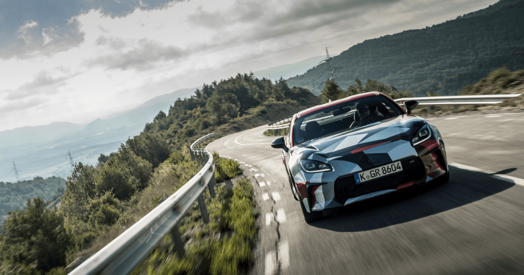 Toyota GR86 rouge et blanche en virage sur une route de montagne sinueuse avec forêts et vallée en arrière-plan