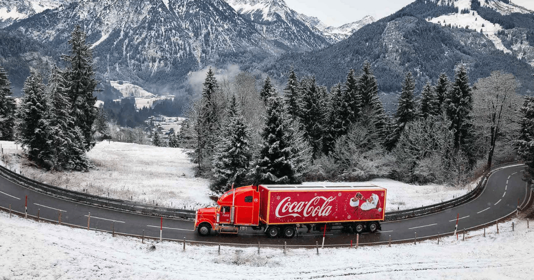 Camion de Noël Coca-Cola rouge et blanc circulant sur une route de montagne enneigée, avec sapins et pics alpins en arrière-plan
