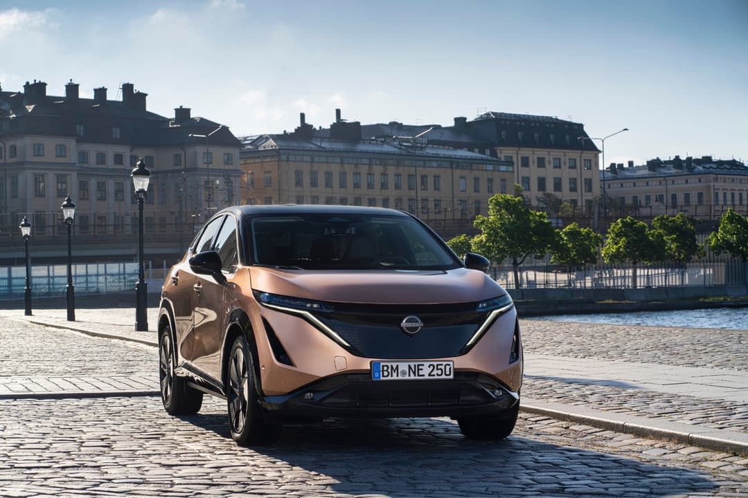Nissan Ariya, SUV électrique beige et noir, garé sur un quai urbain avec immeubles classiques et plan d'eau en arrière-plan