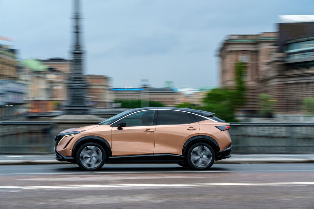 Nissan Ariya couleur bronze en mouvement sur une route urbaine, bâtiments historiques flous en arrière-plan