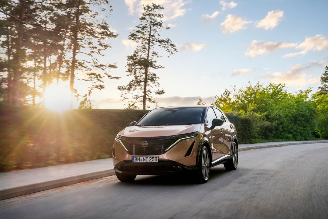 Nissan Ariya blanc en mouvement sur une route de montagne, entouré de pins et de forêt, sous un ciel dégagé avec soleil bas