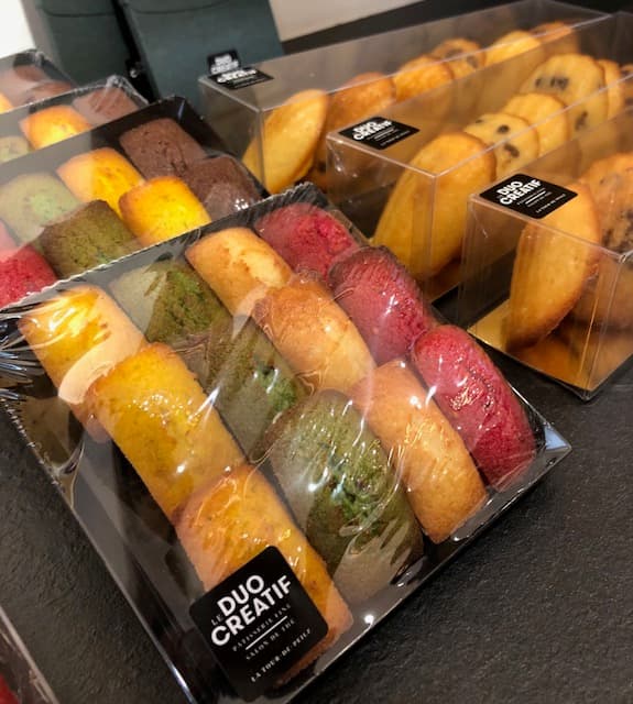 Vitrine de pâtisserie avec madeleines aux saveurs variées : nature, pistache, fruits rouges, présentées en boîtes transparentes étiquetées Duo Créatif