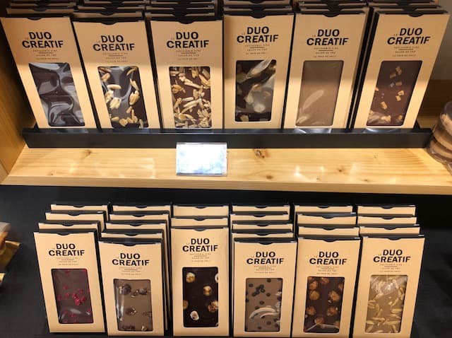 Vitrines de la boutique Duo Créatif présentant des plaques de chocolat et pâtisseries emballées, avec étiquettes beiges et noires, exposées sur des étagères en bois