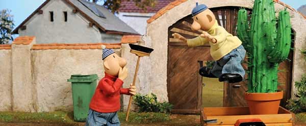 Deux marionnettes enfantines bricolent devant une petite maison décorée, l'une tient une pelle rouge, l'autre grimpe à un cactus vert